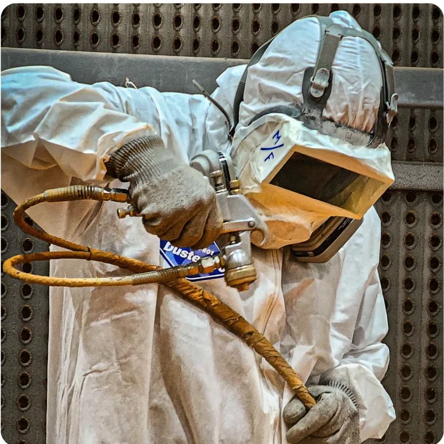 Operator in protective coveralls and respirator spray-painting a metal component with a pneumatic gun